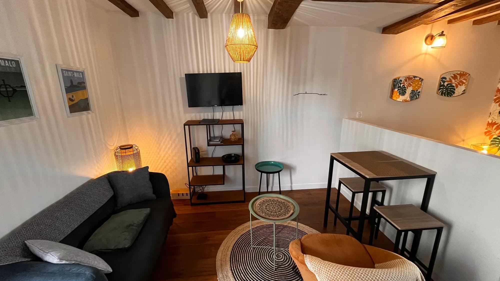 La pièce à vivre du studio Le Secret de Solidor : canapé et coussins sous les poutres anciennes, suspension en rotin, coin repas avec table haute et tabourets, lumière chaude de fin de journée — quartier Solidor à Saint-Servan